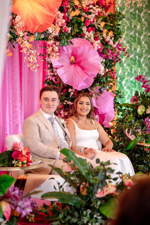 Couple in formal attire posing together surrounded by vibrant pink and floral wedding backdrop decorations