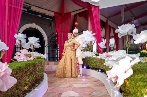 Woman in gold dress standing in ornate courtyard decorated with large white flowers, pink drapes, and green plants