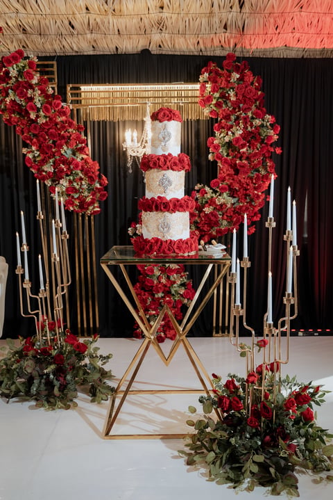 Elegant wedding cake display with three-tiered white cake decorated with red roses, surrounded by red floral installations and gold geometric structures on a white platform