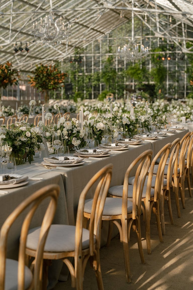 Long banquet table with white floral centerpieces and wooden chairs in a glass greenhouse venue