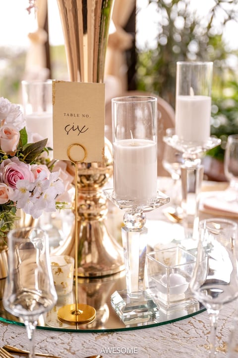 Elegant wedding table centerpiece with pink roses, white candles in glass hurricanes, gold accents, and a kraft table number card on a glass base