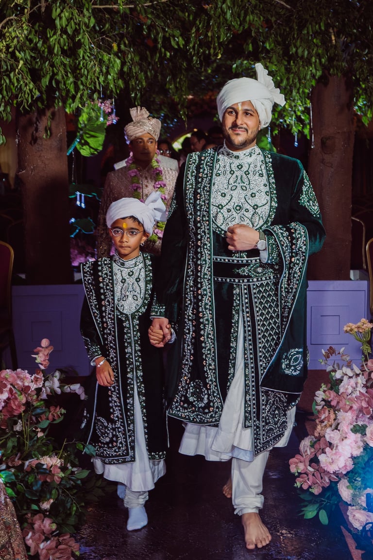 A man and boy wearing matching traditional South Asian wedding attire with white turbans, ornate black and white embroidered jackets, standing together among pink flowers and hanging green foliage.