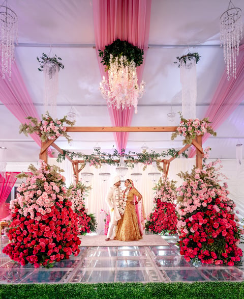 Luxurious wedding ceremony setup with elaborate floral arrangements, pink fabric draping, hanging greenery installations, and bride in gold dress at altar