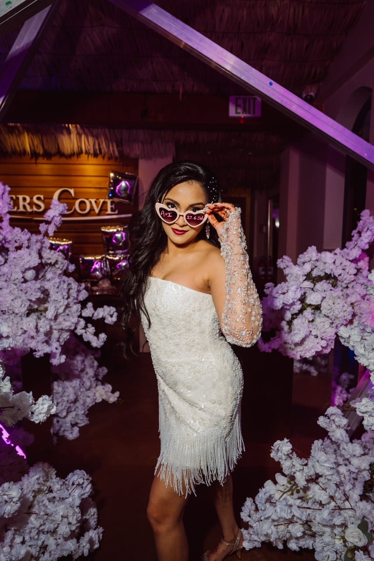 Woman in white sequined dress and sunglasses posing among purple-lit white flowers at an upscale venue