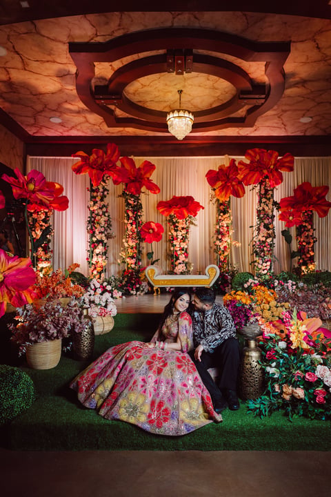 Woman in ornate colorful gown seated in lavishly decorated floral reception with red and gold ceiling and hanging flower installations