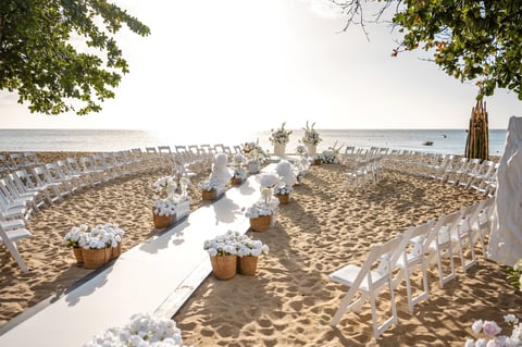 Beach wedding ceremony setup with white chairs lining an aisle decorated with white fabric and lanterns, ocean view in background
