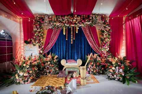 Ornate wedding mandap stage with pink and gold drapes, elaborate floral garland backdrop, and cushioned seating for ceremonial rituals.