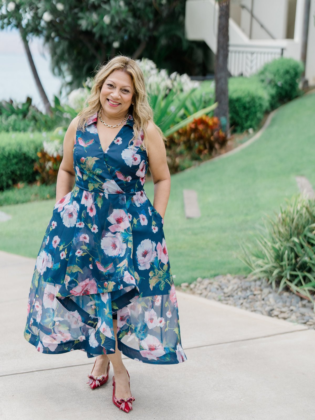 Woman in floral navy dress with blonde wavy hair smiling at camera while standing on a residential driveway lined with landscaping
