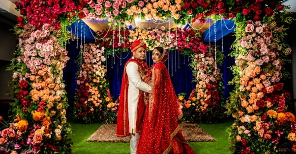 Indian couple in traditional red wedding attire standing under ornate floral arch with colorful roses and hanging decorations