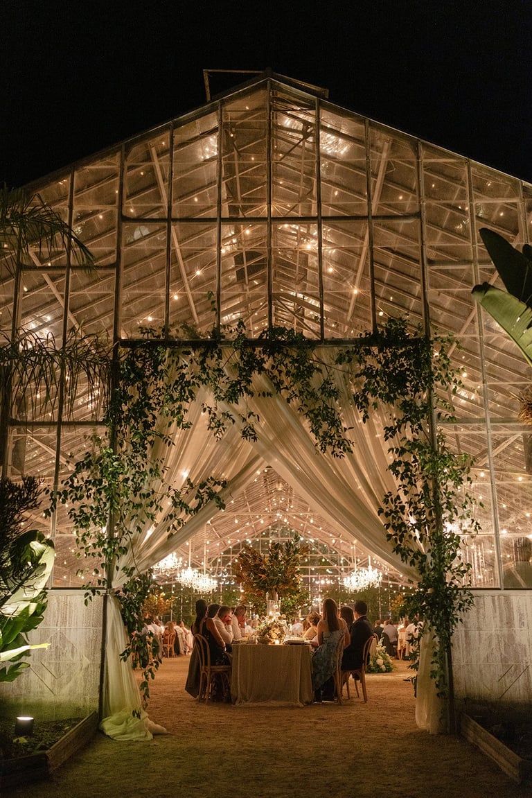 Illuminated glass greenhouse structure at night with ivy vines, draped fabric, and people gathered inside for an elegant event