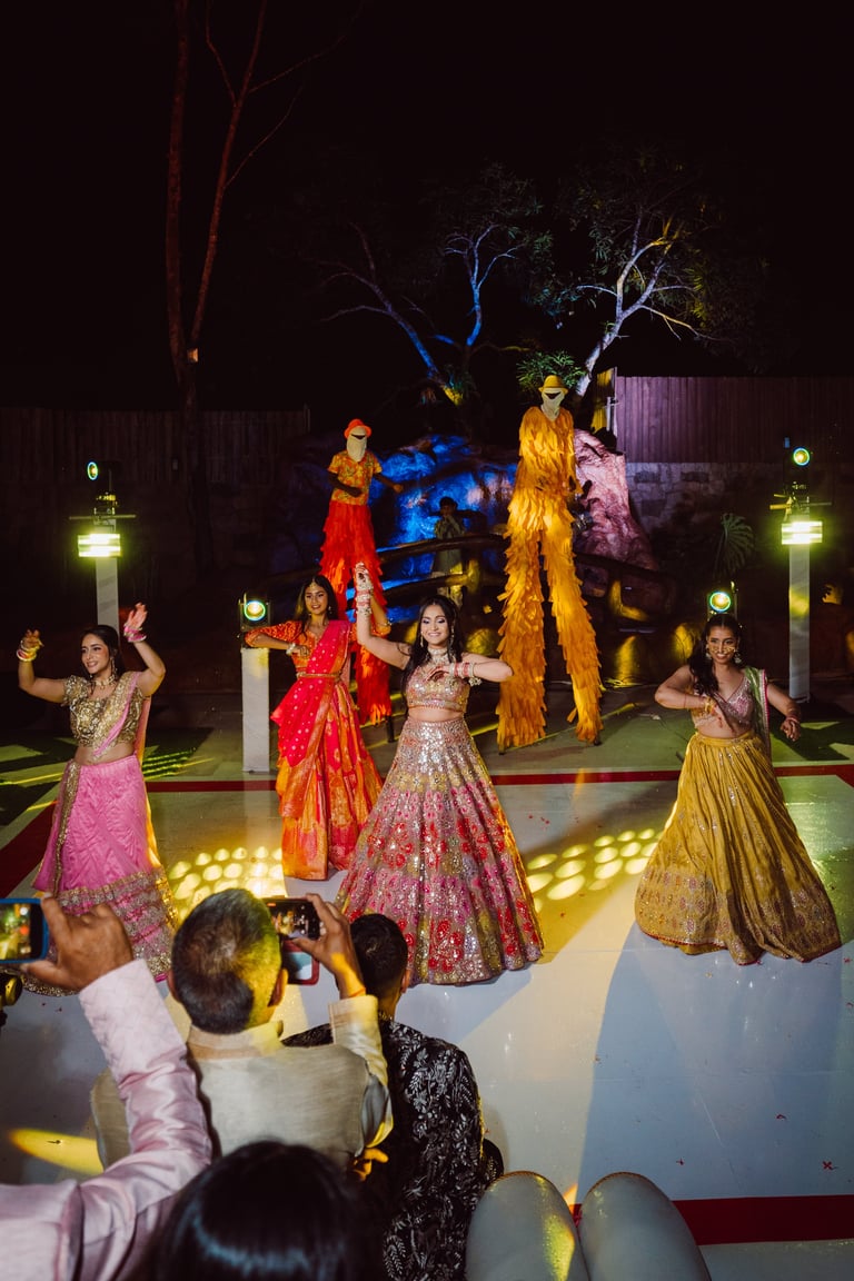 Women in colorful traditional Indian dresses perform on an illuminated stage at night with spectators watching and trees lit in the background