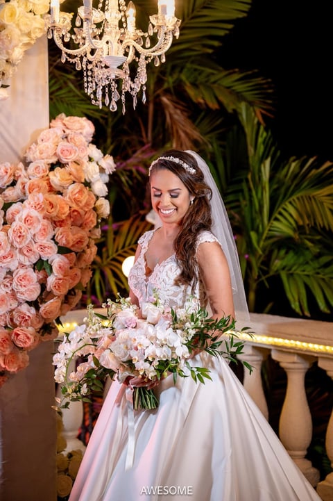 Bride in white lace gown and veil holding bouquet, standing beneath ornate chandelier and pink floral arrangements at elegant wedding venue