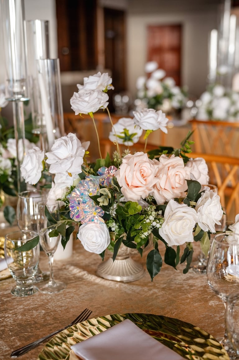 Wedding reception table centerpiece with white and blush roses, glass candleholders, and place settings in an elegant indoor venue.