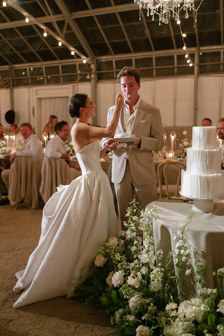 Bride and groom feeding each other cake at wedding reception in elegant venue with white flowers and hanging lights
