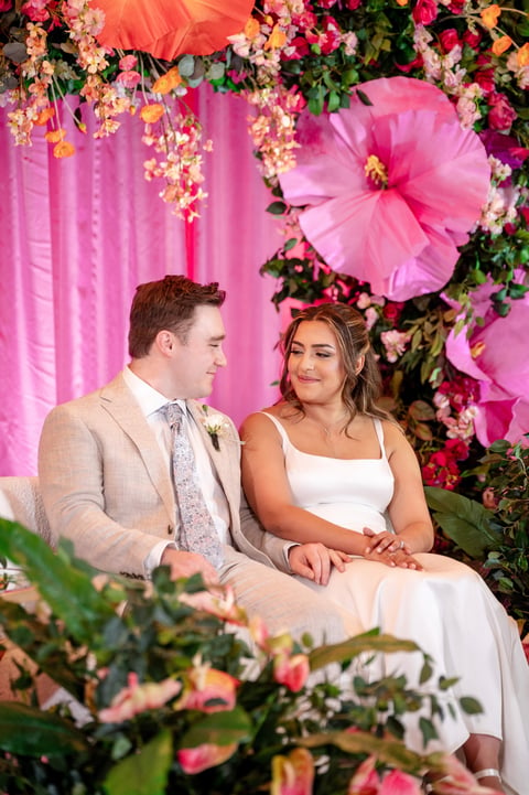 Bride and groom sitting together in front of vibrant pink and floral backdrop with hanging flowers and pink tulle
