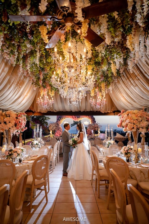 Elegant wedding reception tent with bride and groom in center aisle, hanging floral installations, chandelier, draped fabric ceiling, and decorated tables with flowers