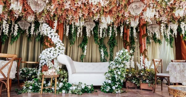 Elegant wedding ceremony setup with white sofa surrounded by lush greenery, hanging floral arrangements, and decorative chairs under draped fabric canopy
