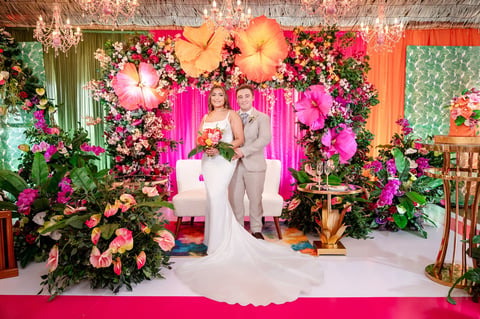 Bride and groom standing under vibrant floral arch with pink, orange, and yellow flowers at wedding ceremony