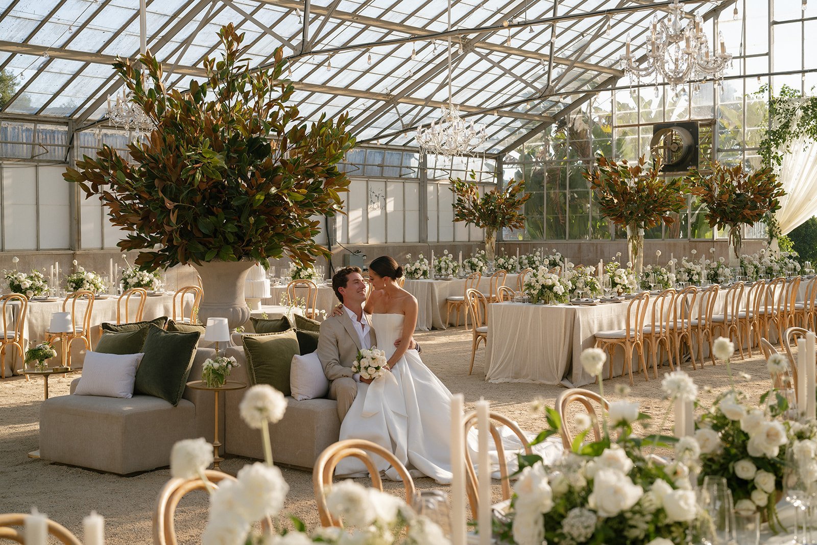Bride and groom sitting on a couch in a glass greenhouse venue decorated with white flowers and greenery for a wedding reception.
