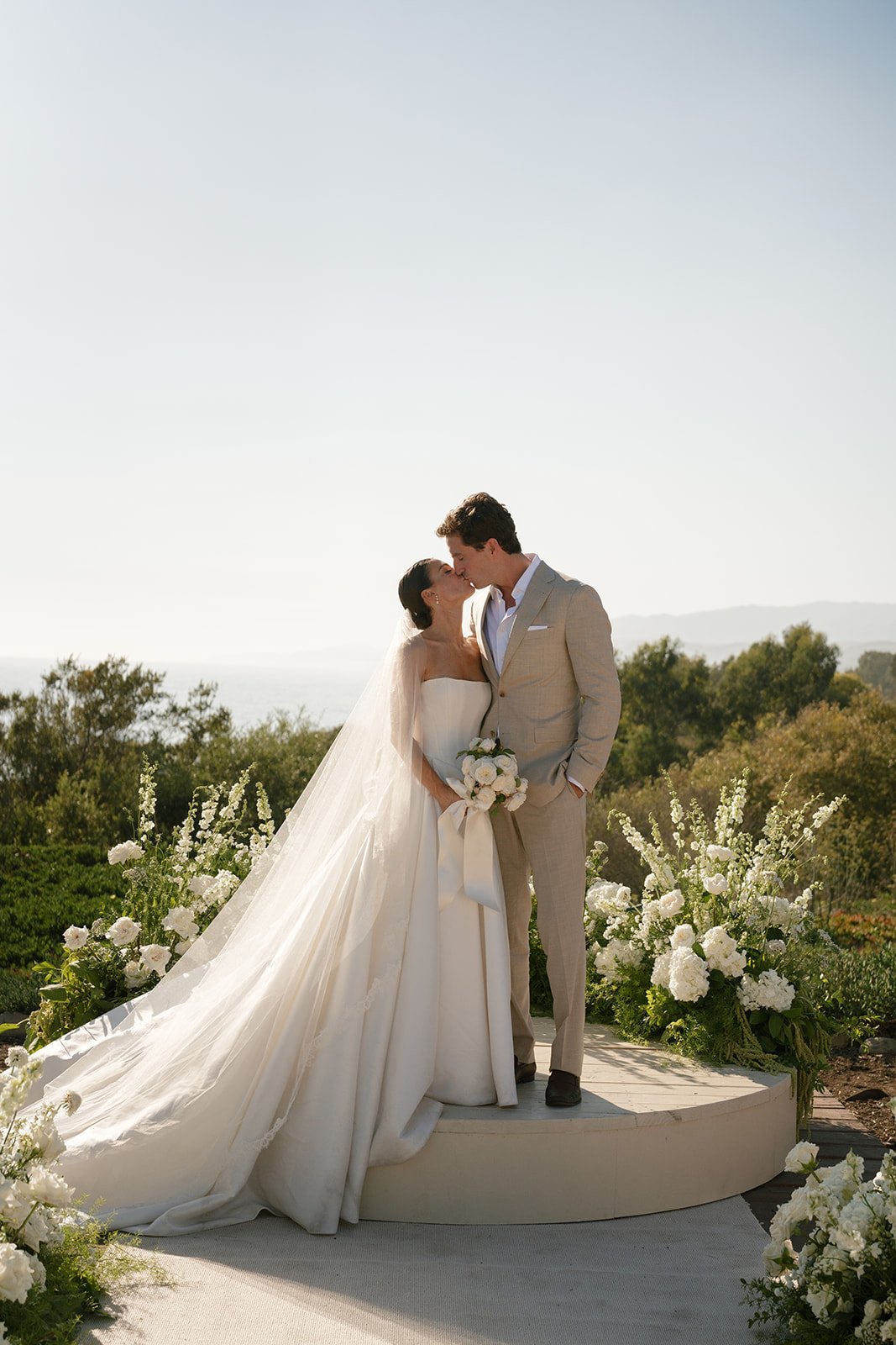 Bride and groom kissing on outdoor wedding platform surrounded by white flowers and mountains in background