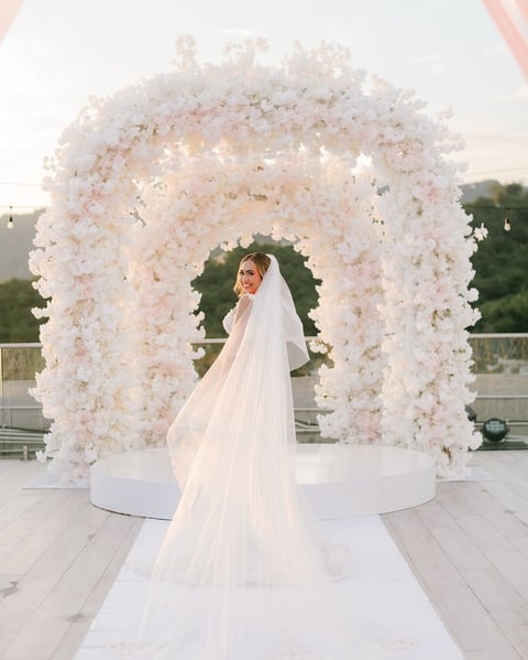 Bride in white wedding dress and veil standing under an elaborate white floral arch on an outdoor terrace
