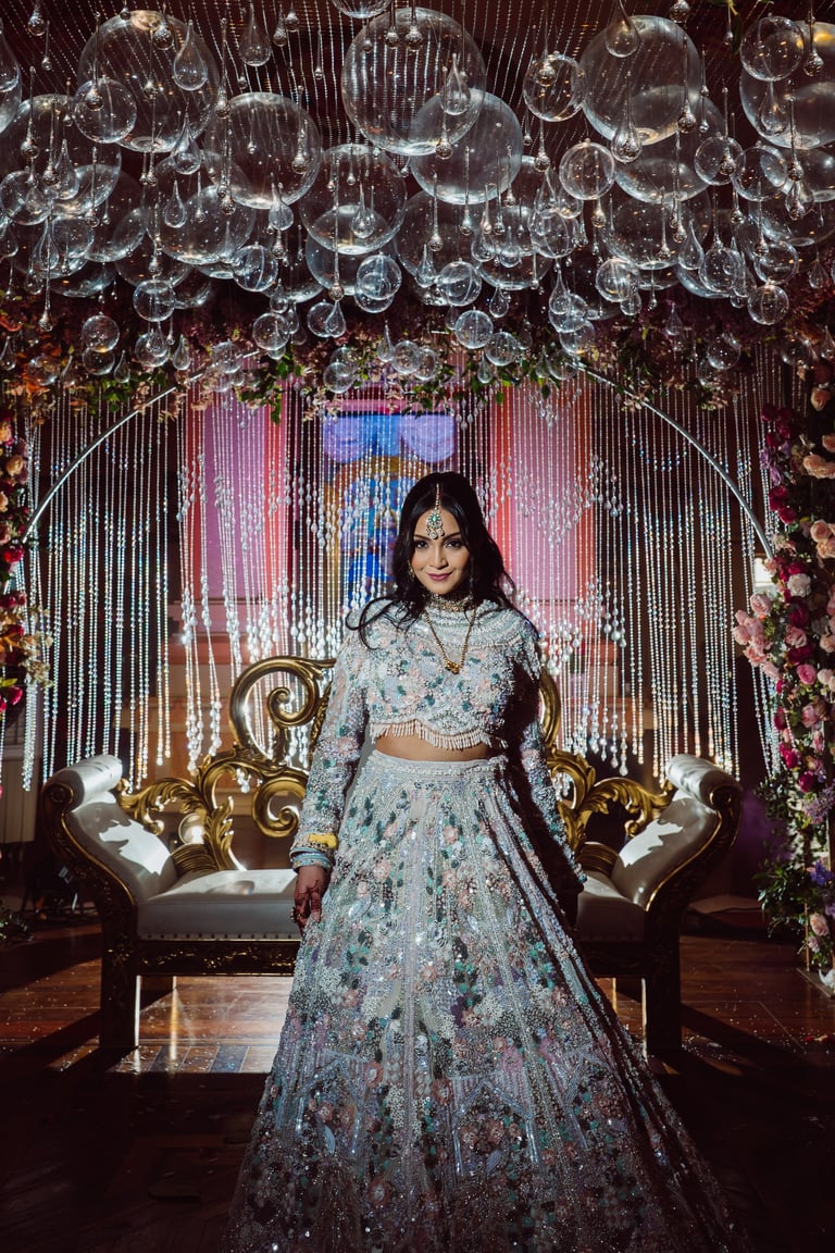 Woman in embellished floral lehenga standing in ornate wedding backdrop with crystal chandeliers and golden furniture