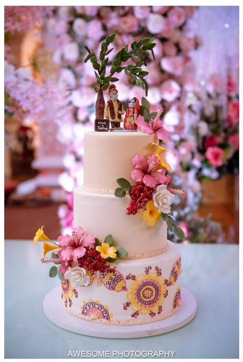 Three-tier white wedding cake decorated with fresh flowers, ornamental patterns, a bamboo plant, and decorative figurines on top