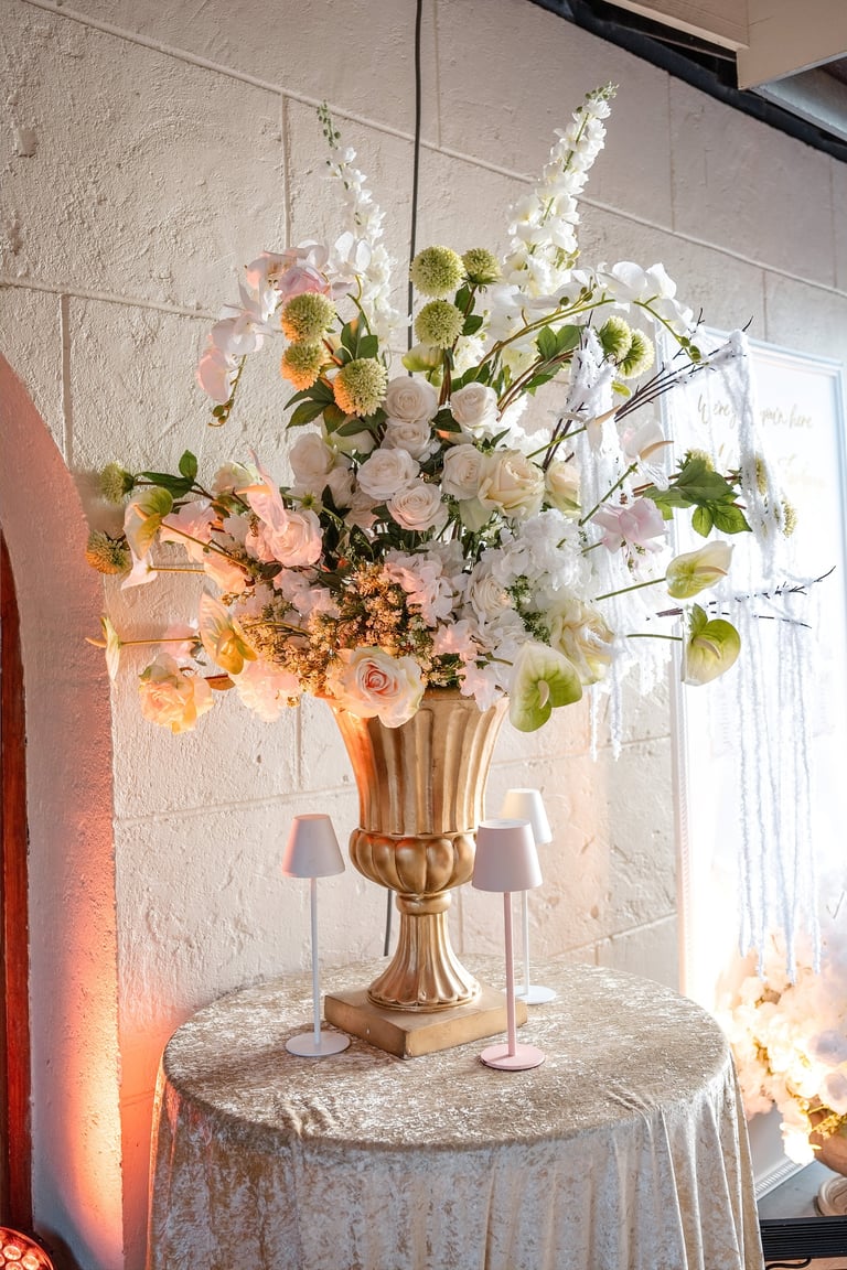 Elaborate floral arrangement in gold urn vase with pink, white, and green flowers on round table with cream tablecloth