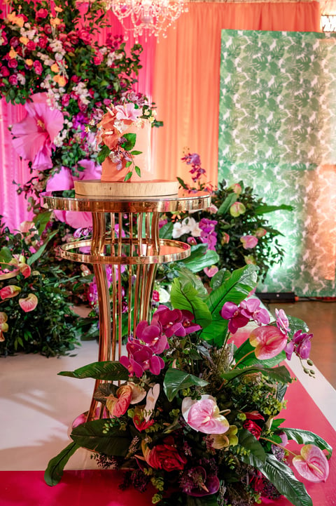 Gold pedestal table surrounded by vibrant pink, red, and white flowers with green foliage against colorful backdrop