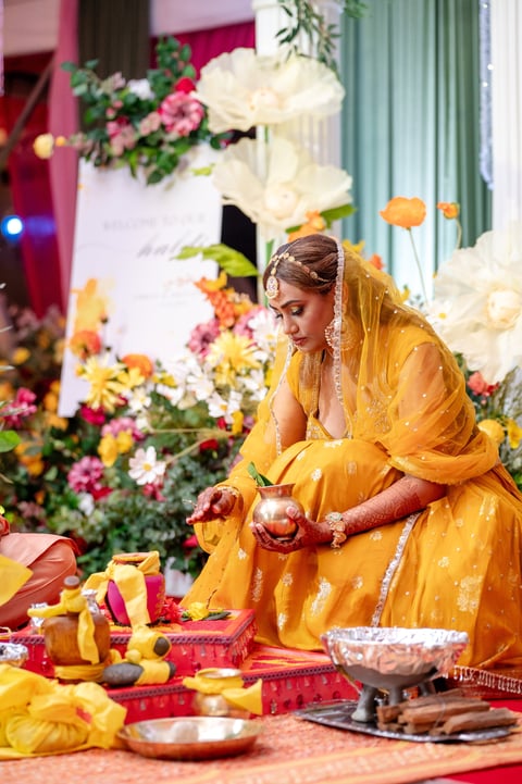 Woman in yellow traditional dress and veil holding pomegranate at ceremonial gathering with colorful flowers and decorative items
