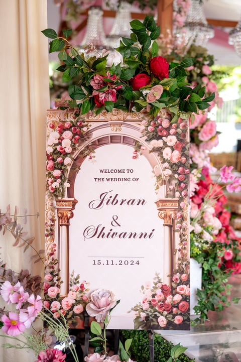 Ornate wedding welcome sign with floral arch displaying Jibran and Shivannis names and date 15.11.2024, surrounded by pink and red roses.