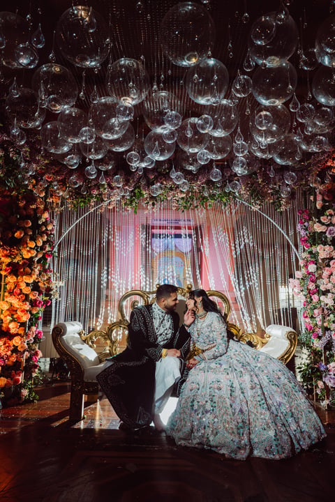 Bride and groom sitting on ornate chairs under elaborate floral and globe lighting installation at wedding reception