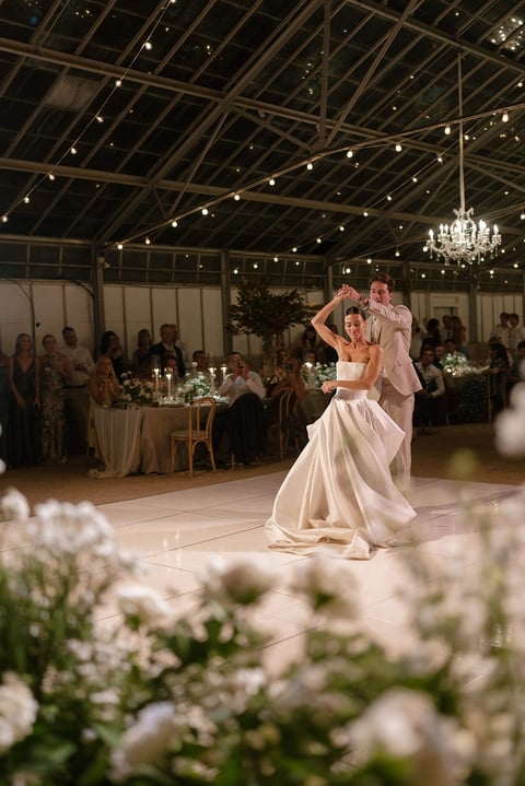 Bride and groom dancing in elegant wedding reception hall with chandelier, white flowers in foreground, and guests at tables in background