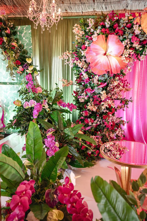 Elaborate floral display with pink and red flowers, green foliage, hanging lights, and large pink petal installation against vibrant pink and green backdrop