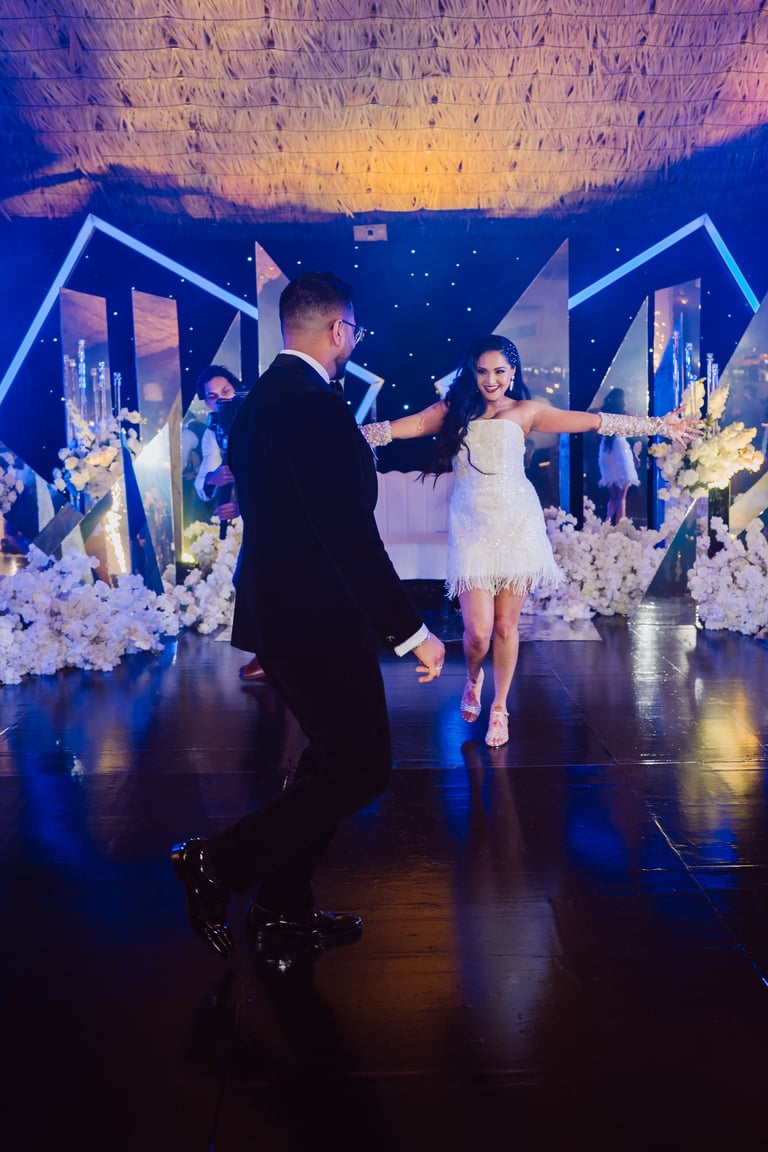 Couple dancing at an elegant wedding reception with blue lighting, white floral arrangements, and decorated backdrop