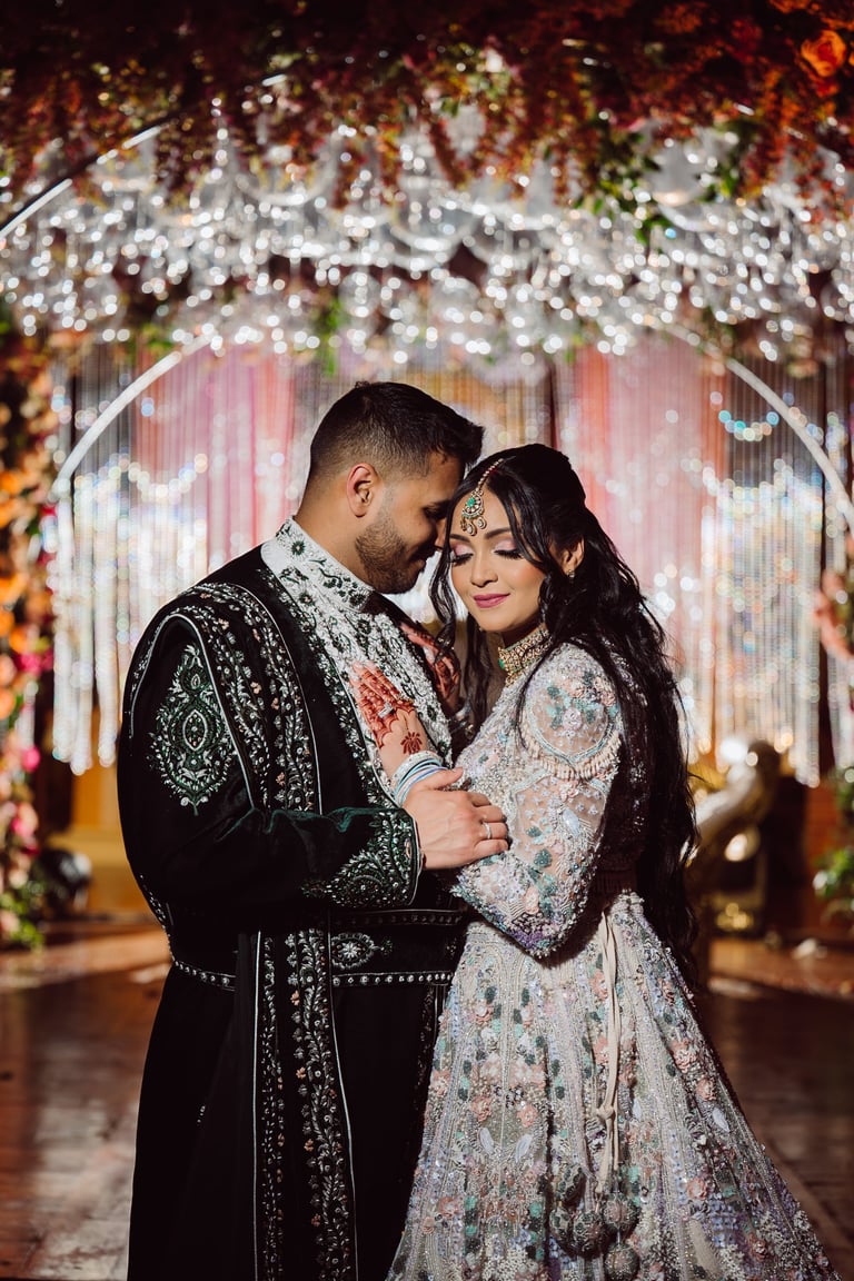 Couple in traditional South Asian wedding attire embracing under an ornate floral arch with golden and white hanging flowers