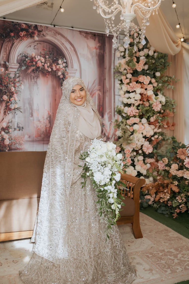 Bride in ornate beaded wedding gown and hijab holding white floral bouquet in decorated event venue with pink and peach flowers