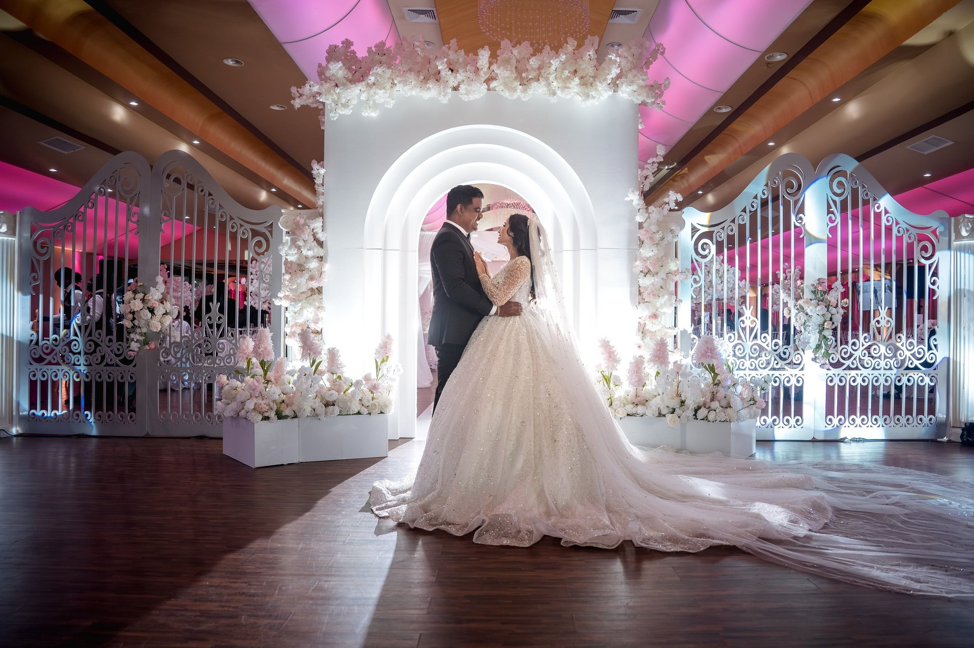 Bride and groom kissing at wedding altar with ornate white arch, pink lighting, and floral decorations in elegant ballroom
