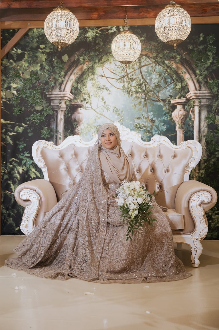 A woman in a beige hijab and embellished gown sits on an ornate white sofa, holding a white flower bouquet, under crystal chandeliers.