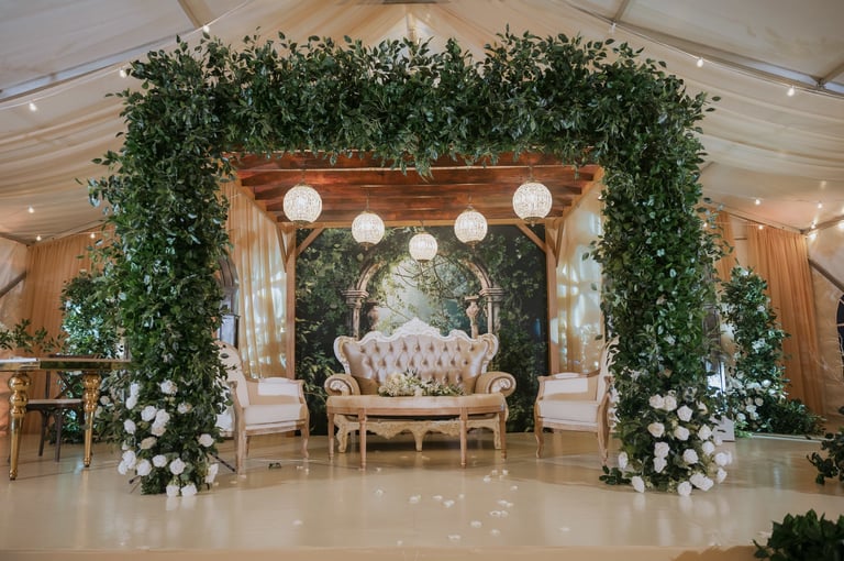 Elegant wedding ceremony backdrop with lush greenery, cream-colored upholstered chairs, hanging lanterns, and white floral arrangements in a decorated tent venue
