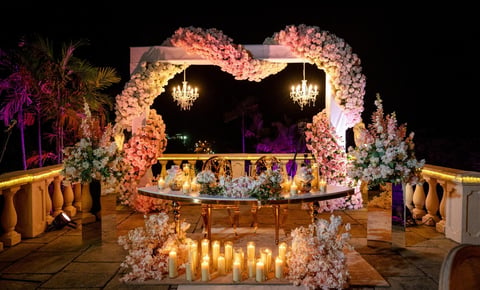 Elegant nighttime wedding ceremony setup with ornate floral arch, glowing candles, chandeliers, and decorated entrance on illuminated rooftop venue