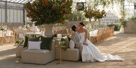 Bride and groom sitting on tan chairs in a glass greenhouse venue with white flowers and wooden chairs visible in the background