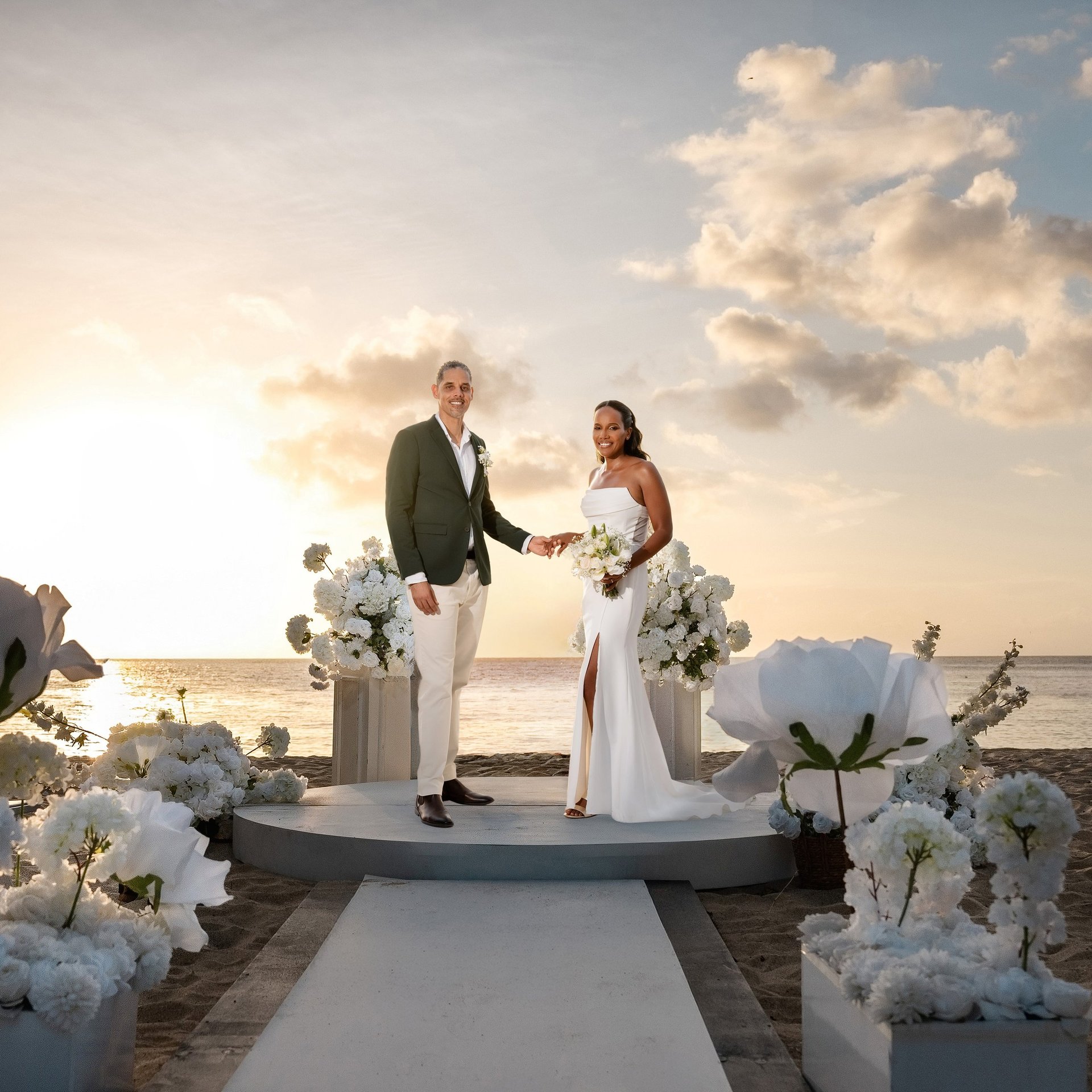 Bride and groom holding hands on a beach platform during sunset ceremony with white floral arrangements and ocean backdrop