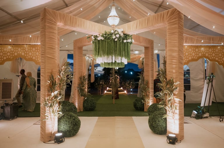 Elegant event entrance with wooden pergola archway, decorated with hanging greenery and floral arrangements, flanked by topiary and ambient lighting