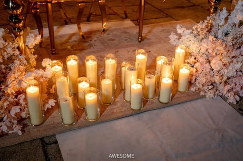 Group of lit candles arranged on floor surrounded by white floral arrangements and wooden chairs in an elegant event space