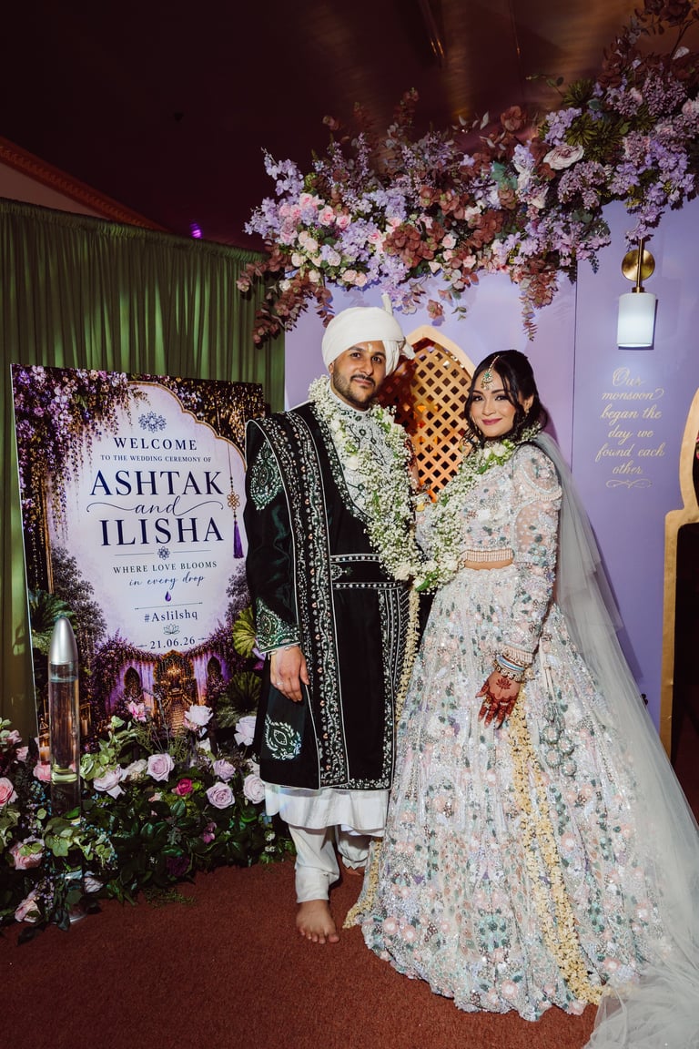 Bride and groom in traditional wedding attire posing together at their reception, with floral decorations and welcome sign behind them