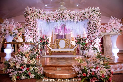 Luxurious wedding reception stage decorated with pink and white floral arrangements, ornate gold furniture, and illuminated backdrop.