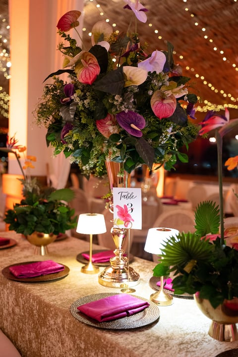 Elegant table setting with tall floral centerpiece featuring pink and purple flowers in gold vase, pink napkins, and warm ambient lighting