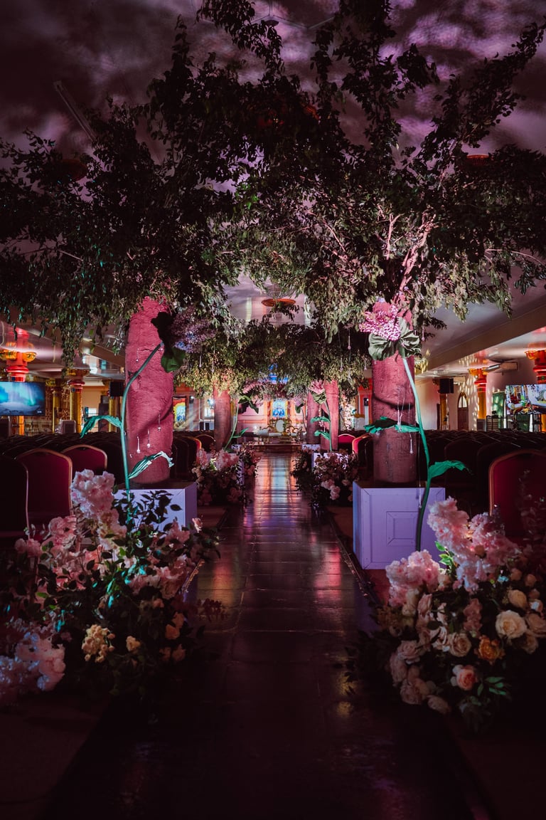 Elegant event venue with lush greenery, pink and white florals, dramatic purple lighting, and a grand floral archway down the center aisle