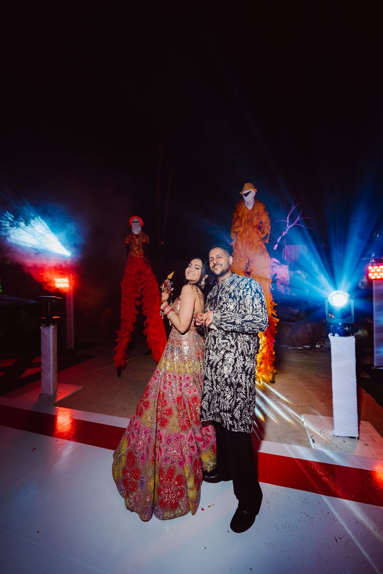 Couple in traditional Indian attire posing under colorful blue and red stage lighting with performers visible in background